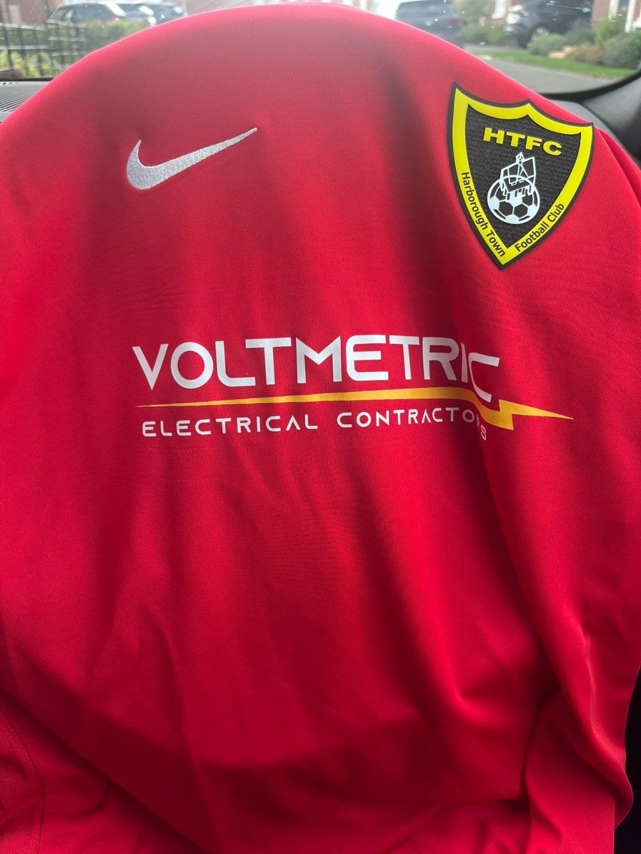 Close-up of the red Harborough Town FC match shirt showing the club badge and the Voltmetric Electrical Contractors sponsor logo.